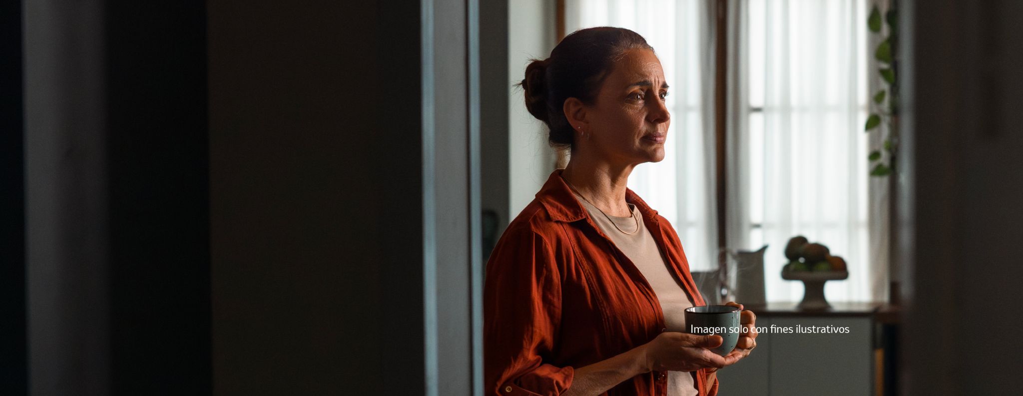A woman with her hair up, wearing a red shirt, holding a cup and gazing thoughtfully out of a window.