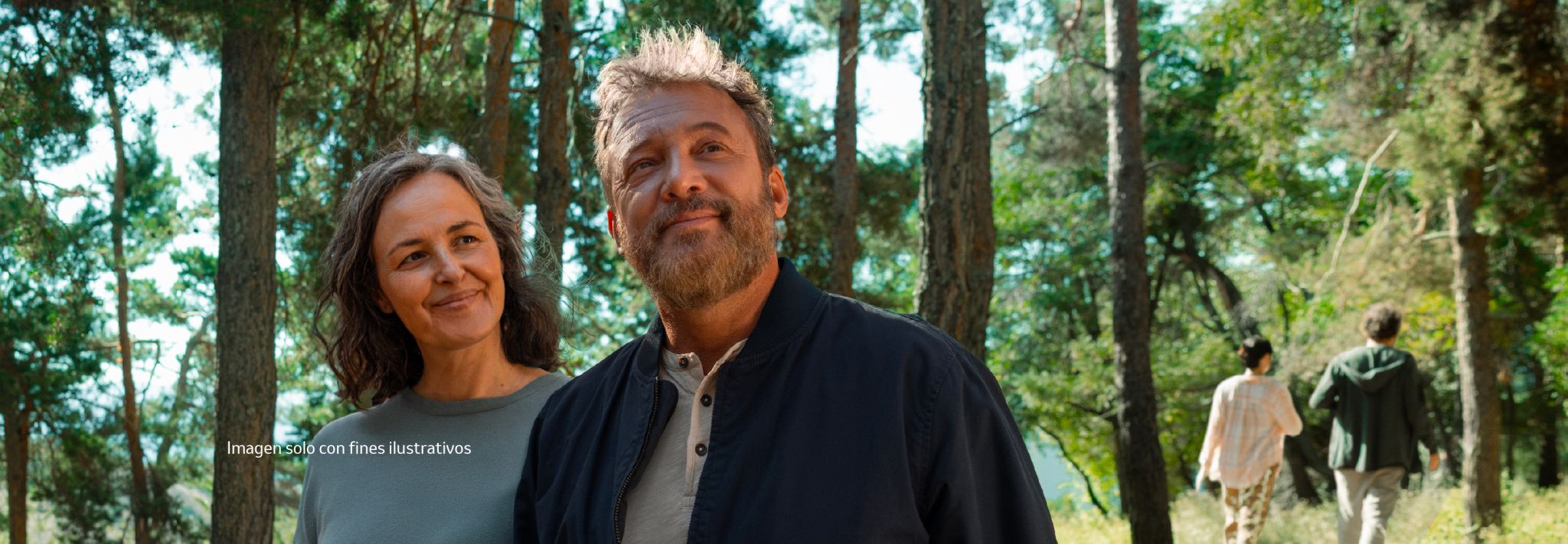 A man and woman smiling gently, looking upward, with tall trees and a soft green background behind them.