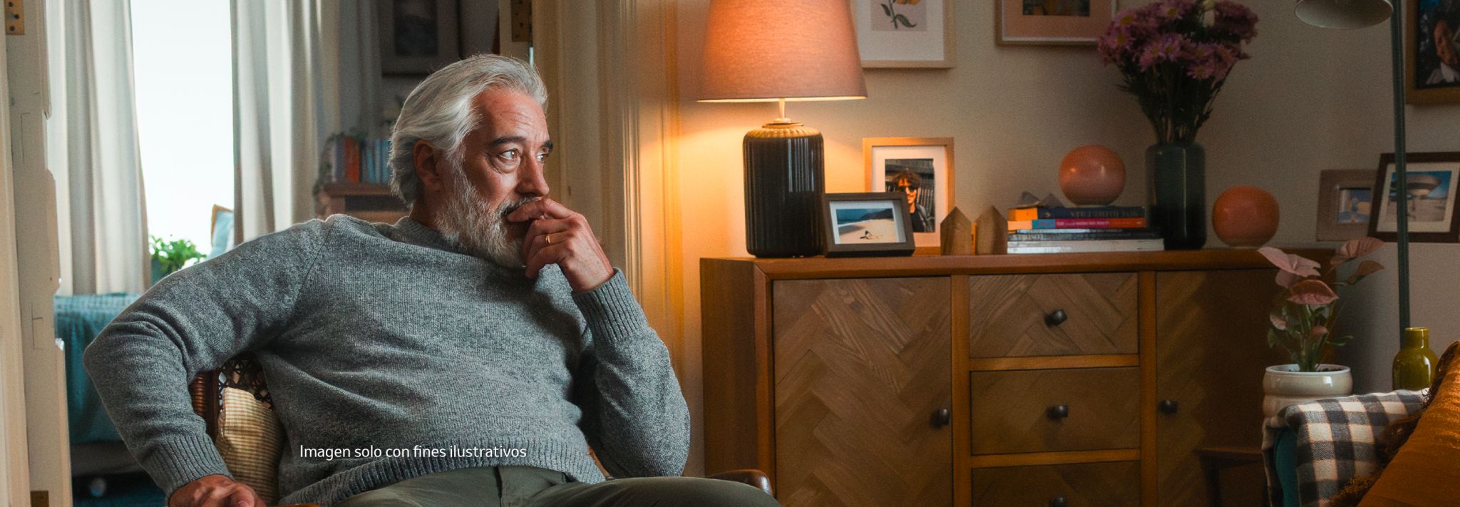 A man with white hair and a beard, wearing a grey sweater, sitting in an armchair with a thoughtful, concerned expression, in a warmly lit living room.